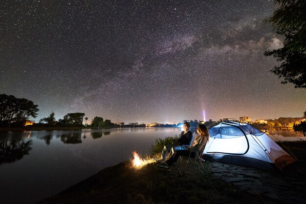 Notte in campeggio sulla riva del lago. Uomo e donna che si siedono sulle sedie vicino al falò e alla tenda luminosa, godendo della vista del cielo serale pieno di stelle e via lattea, superficie dell'acqua tranquilla, città luminosa