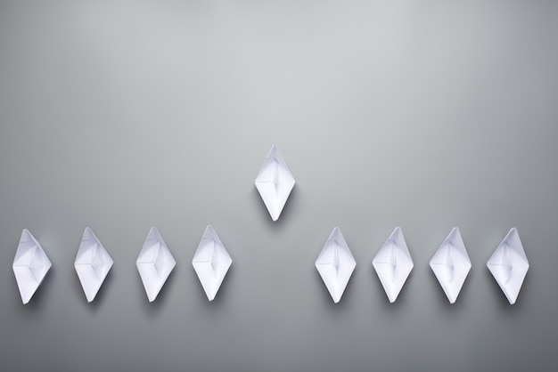 Nove barche origami fatte di carta su sfondo grigio con una che guida l'altra in un'immagine concettuale.