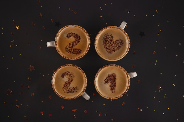Numeri di Capodanno su tazze di caffè schiumate scritte da polvere di cacao su sfondo nero tra stelle dorate