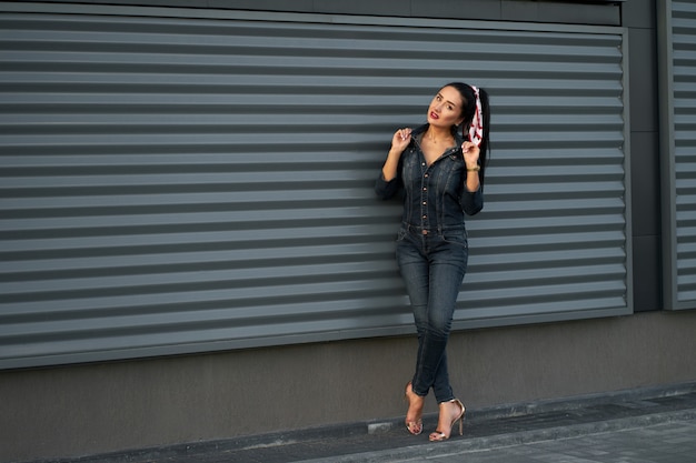 Nuova collezione alla moda. Ragazza castana alla moda abbastanza giovane del ritratto con capelli lunghi e le labbra rosse che indossano la tuta moderna del denim sulla parete urbana grigia