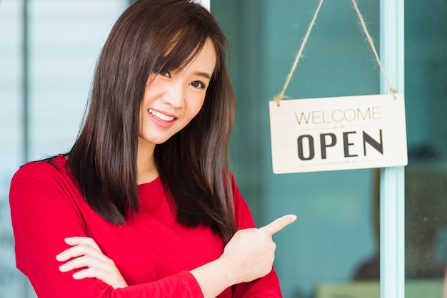 Nuova normale, giovane donna asiatica felice e sorridente punta il dito per notare l'etichetta della tavola di legno "WELCOME OPEN" appesa attraverso la porta di vetro del negozio, l'attività si apre dopo la pandemia di coronavirus