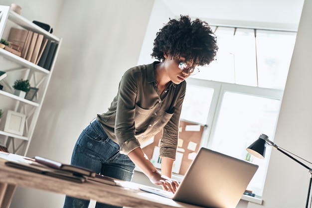 Nuova soluzione ogni giorno. Attraente giovane donna africana che usa il laptop mentre lavora in ufficio