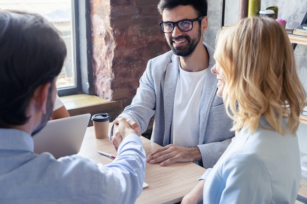 Nuovi partner commerciali. Giovani colleghi moderni in abbigliamento casual intelligente che si stringono la mano e sorridono mentre sono seduti nell'ufficio creativo.