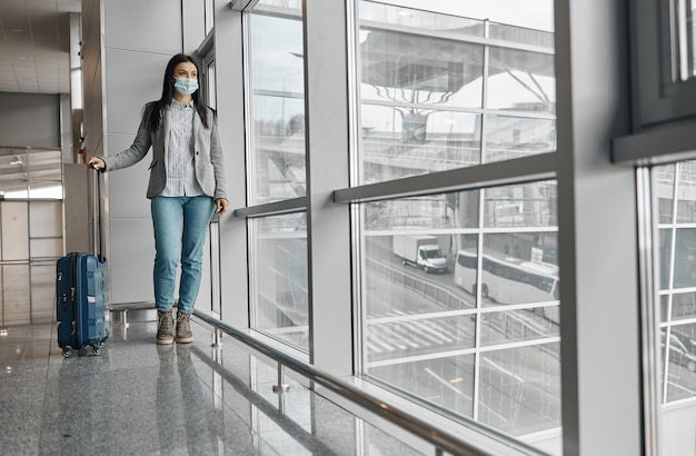 Nuovo ordine negli aeroporti. Donna in maschera nel terminal con bagagli vicino a finestre panoramiche da sola