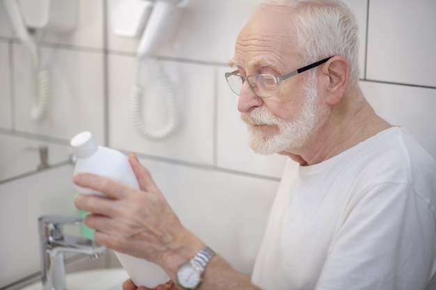 Nuovo shampoo. Uomo dai capelli grigi in occhiali che esamina la bottiglia di shampoo