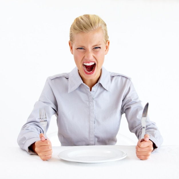 Nutrimi ora Giovane donna furiosa seduta davanti a un piatto vuoto mentre tiene in mano coltello e forchetta