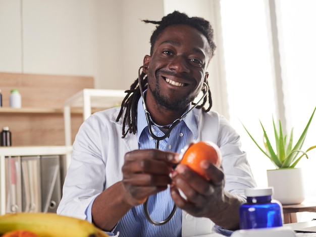 Nutrizionista afroamericano guardando la fotocamera e mostrando frutti sani nella consultazione