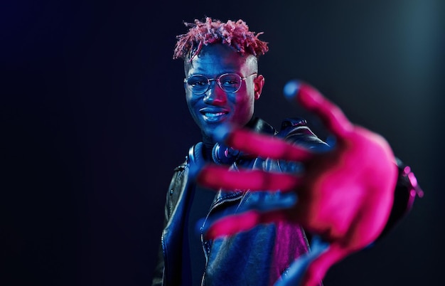 Occhiali alla moda e cuffie wireless Illuminazione al neon futuristica Giovane uomo afroamericano in studio