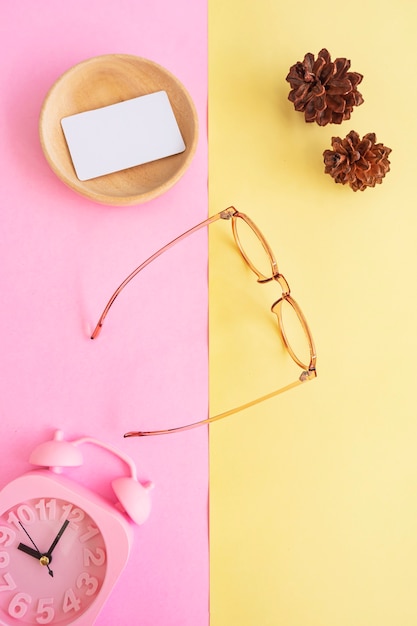 Occhiali ovali nella foto in stile estivo minimale su sfondo rosa pastello e giallo. Sveglia, fiori di pino, biglietti da visita