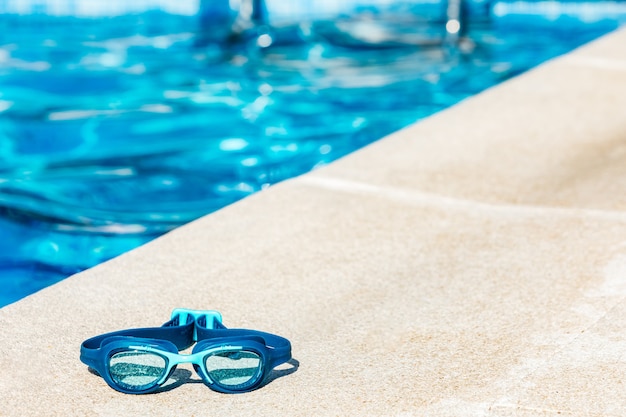 Occhialini da nuoto blu sul bordo della piscina, nell'angolo in basso a sinistra, con l'acqua blu in lontananza
