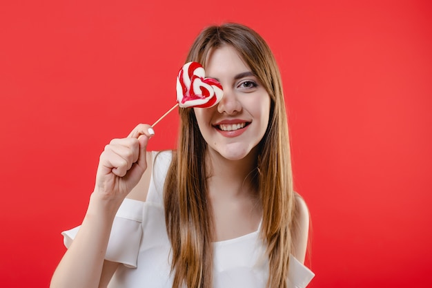 Occhio della copertura della donna con la camicetta bianca d'uso della caramella del lollypop a forma di cuore isolata sulla parete rossa