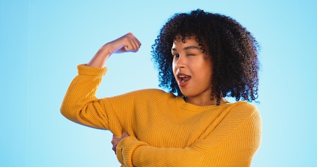 Occhiolino braccio e bicipite con una donna di colore che scherza in studio su uno sfondo blu per divertimento o umorismo Giocoso forte e muscoloso con un'attraente giovane donna comica che si flette mentre strizza l'occhio o ride