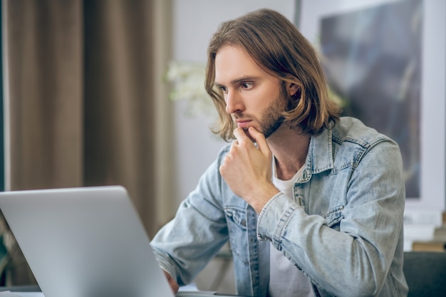 Occupato con il lavoro. Uomo dai capelli lunghi in camicia di jeans che lavora al computer portatile e che sembra concentrato