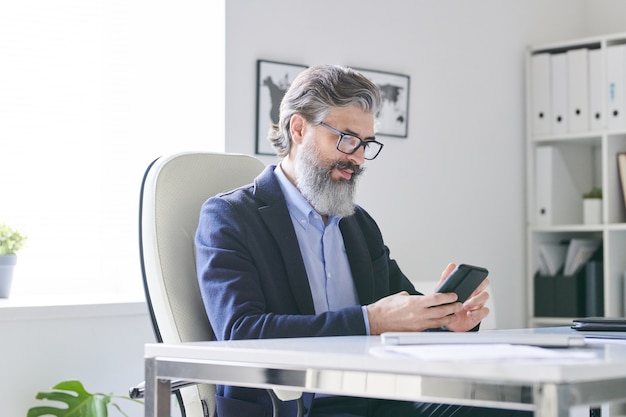 Occupato professionista senior con i capelli grigi e la barba che scorre nello smartphone mentre controlla l'orario dei suoi clienti o candidati in ufficio