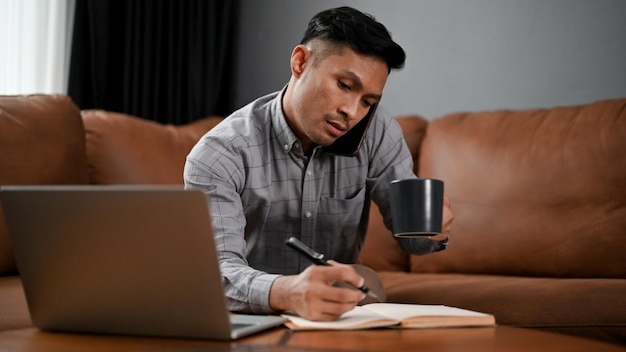 Occupato uomo d'affari asiatico maturo sorseggiando caffè e prendendo appunti mentre si parla al telefono