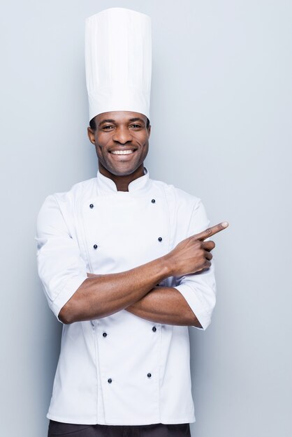 Offerta speciale dello chef. Fiducioso giovane chef africano in uniforme bianca rivolto lontano e sorridente
