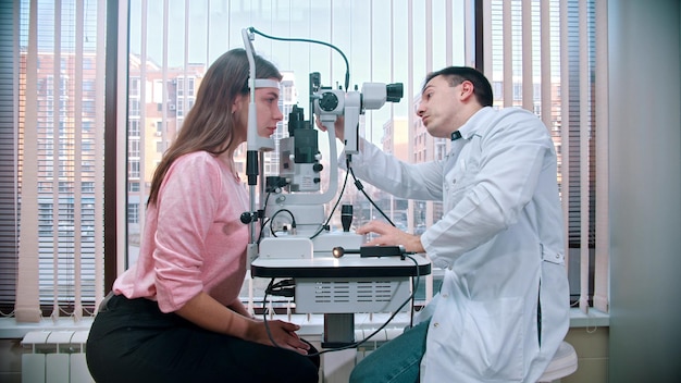 Oftalmologia un medico che controlla l'acuità visiva della giovane donna graziosa con un'attrezzatura speciale nell'armadio spazioso sullo sfondo della finestra panoramica