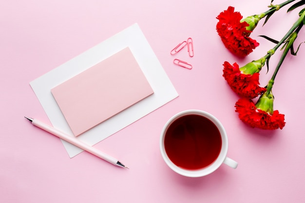 Oggetti rosso-rosa. Tazza di tè, fiori di garofano Blocco note per testo su sfondo rosa pastello.