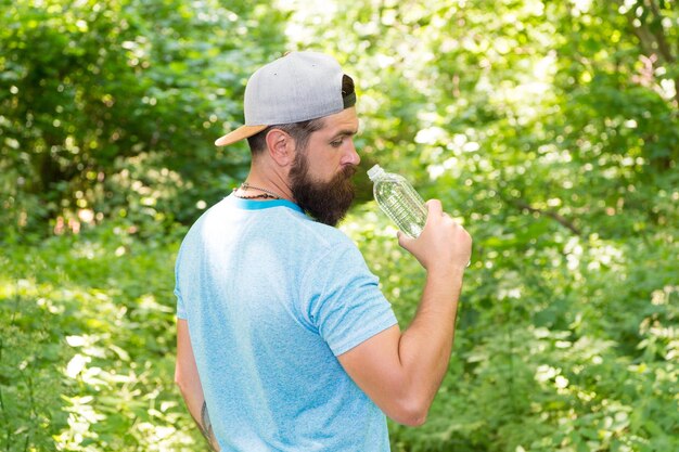 Ogni sorso conta ragazzo maturo in cappello viaggiatore assetato hipster acqua potabile nella foresta all'aperto con tramonto natura sullo sfondo Uomo barbuto con bottiglia d'acqua fitness ritratto di uomo barbuto