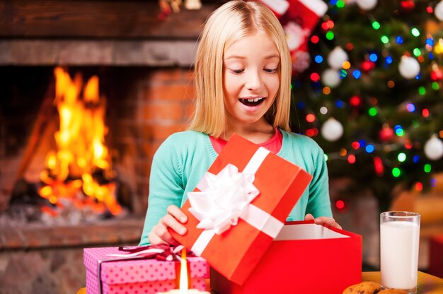 Oh! Bambina carina che apre una scatola regalo e sorride mentre è seduta al tavolo con l'albero di Natale e il caminetto sullo sfondo