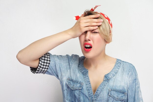 Oh ho perso tutto. Ritratto di triste perdente di giovane donna in casual camicia di jeans blu con fascia rossa in piedi tenendole la testa, preoccupata, sentendosi male. foto in studio al coperto, isolata su sfondo bianco