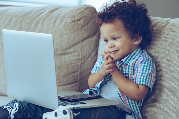 Oh i miei cartoni preferiti online! Piccolo neonato africano che sorride e guarda il laptop mentre è seduto sul divano di casa