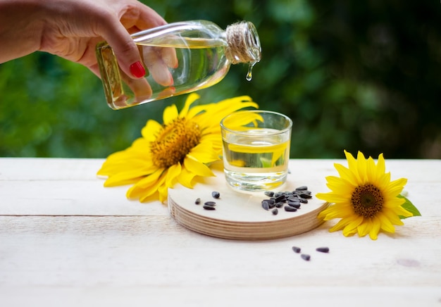 Olio di girasole che versa dalla bottiglia in un bicchiere con girasoli e semi sullo sfondo