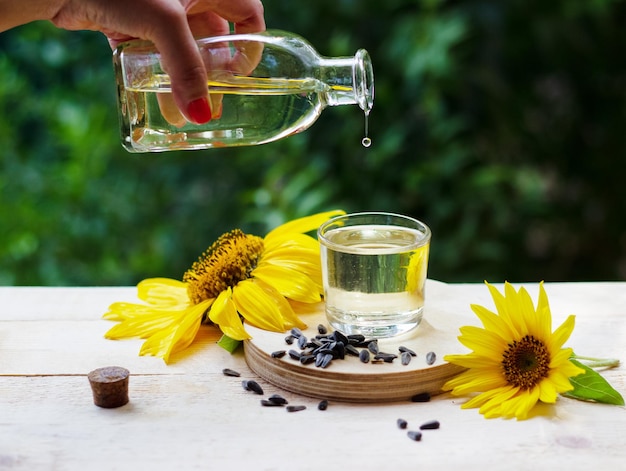 Olio di girasole che versa dalla bottiglia in un bicchiere con girasoli e semi sullo sfondo