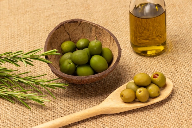 Olive verdi in cucchiaio di legno e in guscio di cocco Olio d'oliva in bottiglia e un rametto di rosmarino