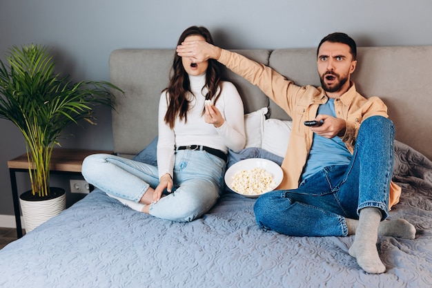 Omg che cos'è La dolce coppia ha il tempo libero in quarantena guarda il film impressionato sorpreso inaspettato film thriller che finisce coprendosi gli occhi con una scatola di pop corn sedersi sul divano in casa al chiuso Bella coppia guarda