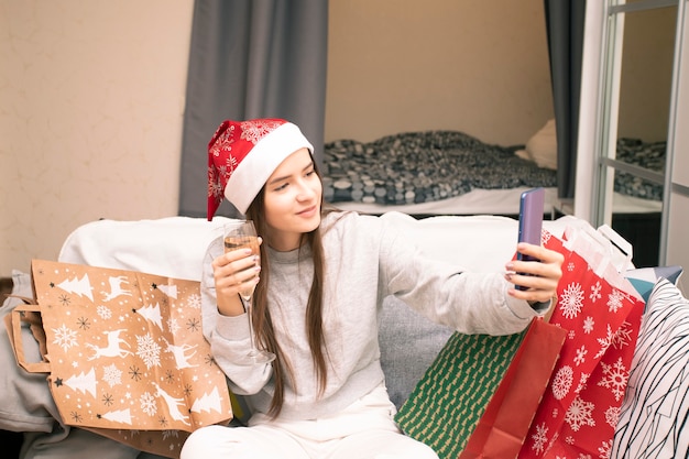 Online Capodanno e auguri di Natale. Una ragazza con un cappello da Babbo Natale chiama la famiglia e gli amici tramite collegamento video con un bicchiere di champagne