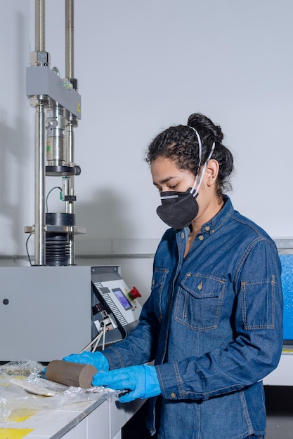 Operaia industriale femminile con maschera protettiva e guanti in lattice blu in un laboratorio di prove geologiche che disimballa un cilindro di argilla