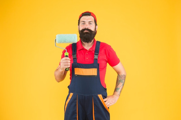 Operaio di riparazione domestica uomo barbuto lavoratore con strumento rullo hipster builder in cappuccio sfondo giallo erettore assistente in uniforme da lavoro ingegnere progettista pronto a dipingere il concetto di riparazione della parete