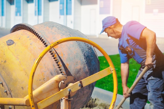 Operaio edile in uniforme lavora in cantiere il giorno d'estate Il muratore anziano spala cemento e sabbia nella betoniera Flusso di lavoro autentico