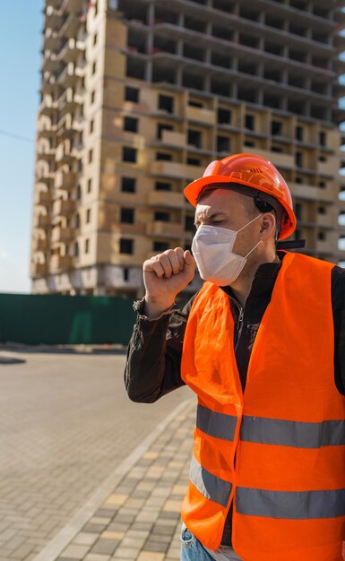 Operaio edile maschile in tuta e maschera medica che tosse sullo sfondo della casa in costruzione Concetto di minaccia di infezione