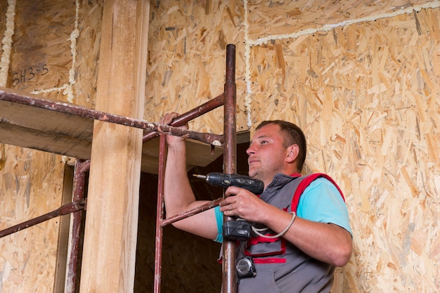 Operaio edile maschio costruttore tenendo trapano cordless salendo la scala di impalcature all'interno di casa incompiuta con telaio di pannelli di compensato di particelle esposte