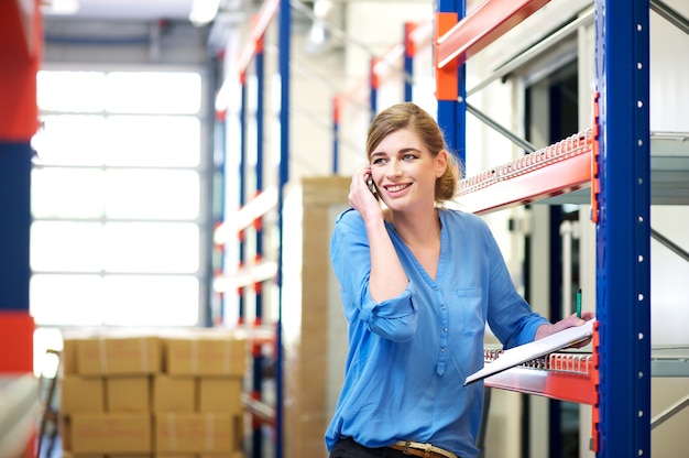 Operaio femminile di logistica che controlla azione e che parla sul cellulare in magazzino