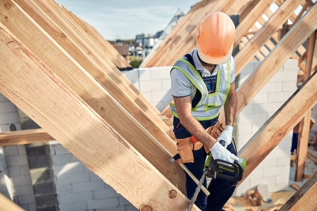 Operaio impegnato in un cantiere che indossa dispositivi di protezione e utilizza una motosega per tagliare il legno per la costruzione