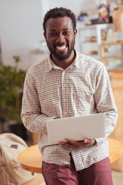 Operaio più felice. Gioioso giovane uomo seduto sul tavolo in caffè, che tiene un computer portatile e sorride allegramente alla telecamera