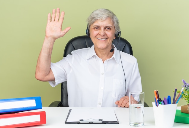 Operatore di call center femminile caucasico sorridente sulle cuffie seduto alla scrivania con strumenti da ufficio alzando la mano isolata sul muro verde