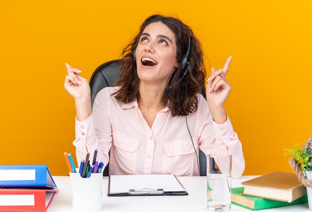 Operatore di call center femminile piuttosto caucasico sorpreso sulle cuffie seduto alla scrivania con strumenti da ufficio che guardano e puntano verso l'alto isolato sul muro arancione