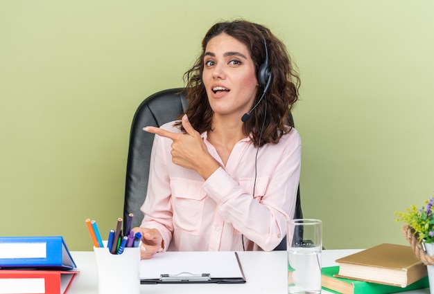 Operatore di call center femminile piuttosto caucasico sorpreso sulle cuffie seduto alla scrivania con strumenti da ufficio che puntano a lato isolato sul muro verde