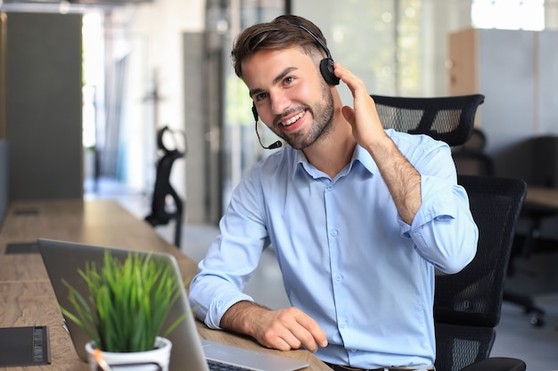 Operatore di call center maschio sorridente con cuffie seduto in un ufficio moderno, consultando informazioni online in un laptop, cercando informazioni in un file per essere di aiuto al cliente.