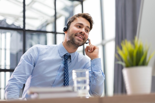 Operatore maschio del telefono di supporto giovane allegro in cuffia sul posto di lavoro con i colleghi sullo sfondo mentre si utilizza il servizio di assistenza del computer e il concetto di call center di consulenza clienti