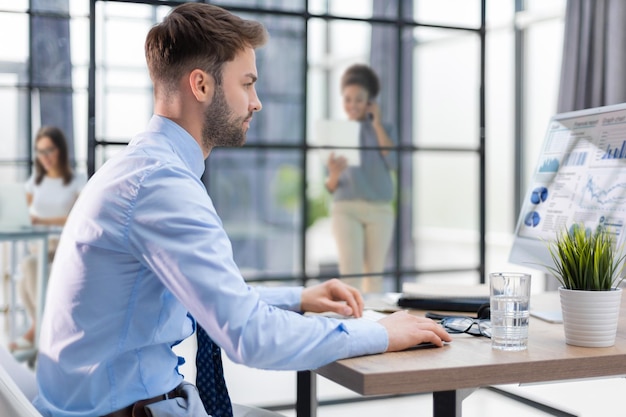 Operatore maschio del telefono di supporto giovane allegro in cuffia sul posto di lavoro con i colleghi sullo sfondo mentre si utilizza il servizio di assistenza del computer e il concetto di call center di consulenza clienti