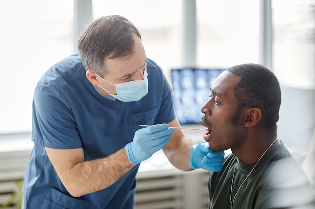 Operatore medico che indossa maschera protettiva e guanti che prendono un tampone orale da un giovane soldato nero per pcr