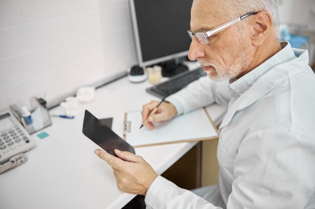 Operatore sanitario anziano concentrato che indossa occhiali e guarda il tablet mentre prende appunti