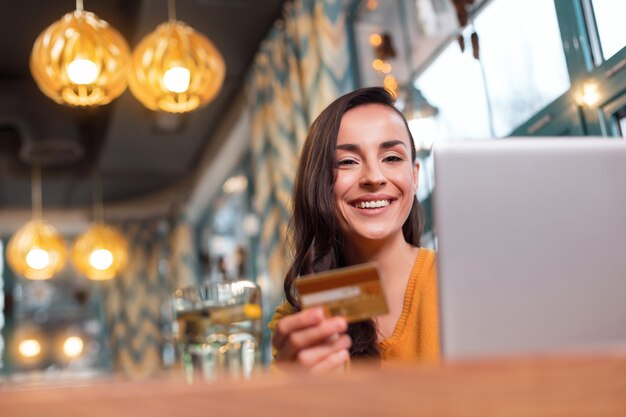 Opportunità di pagamento. Basso angolo di positiva giovane donna bruna che trasportano carta di credito mentre sorridendo e utilizzando laptop