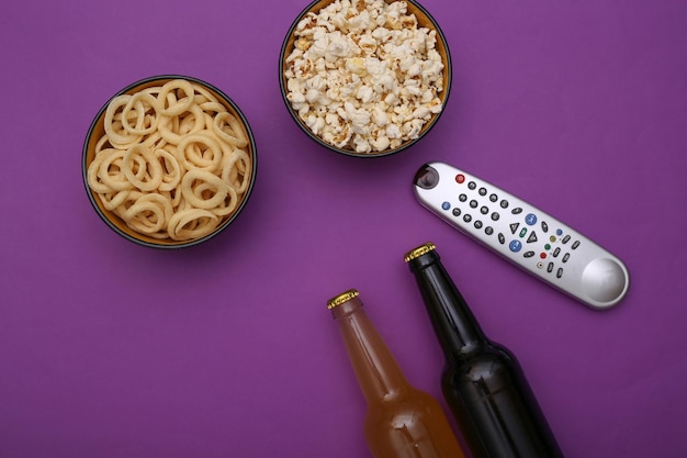 Ora della TV. Bottiglie di birra, telecomando della tv e ciotola con snack su sfondo viola. Vista dall'alto