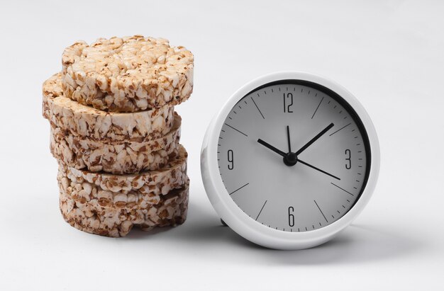 Ora di colazione. Concetto di dieta. Pila di pane croccante di grano intero e orologio sulla superficie bianca